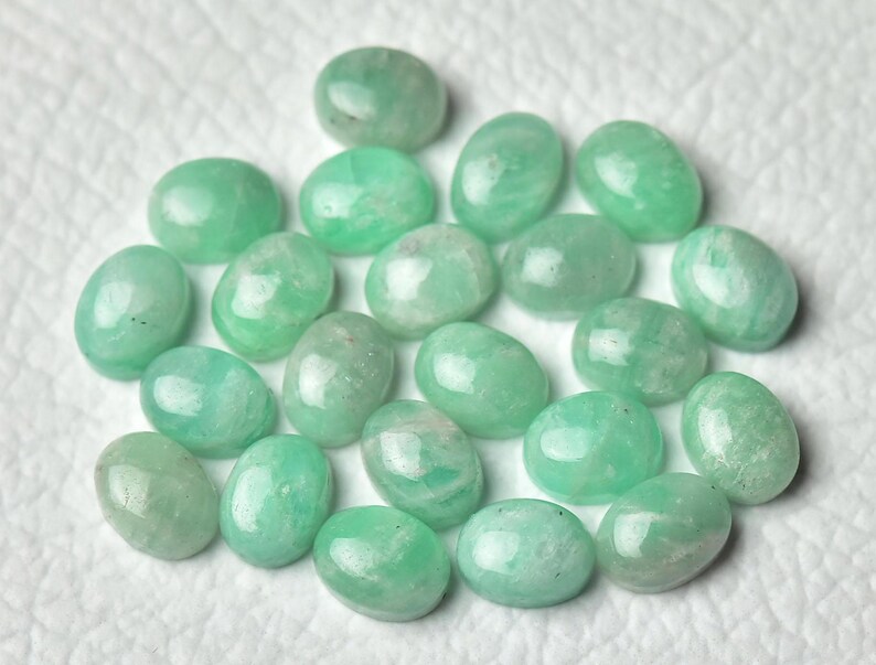 Peut inclure: Une collection de 20 pierres pr&eacute;cieuses vertes de forme ovale, probablement de l'amazonite, avec une finition lisse et polie. Les pierres sont dispos&eacute;es de mani&egrave;re al&eacute;atoire sur une surface blanche.