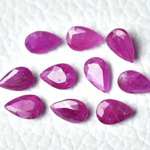Op de afbeelding: Tien peervormige roze edelstenen, waarschijnlijk robijnen, zijn gerangschikt op een wit, getextureerd oppervlak. De edelstenen zijn gefacetteerd en hebben een glanzende afwerking.