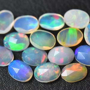 Könnte beinhalten: Eine Sammlung ovaler Opale in verschiedenen Schattierungen von Weiß, Blau, Grün und Rosa. Die Edelsteine sind auf einer schwarzen Oberfläche angeordnet.