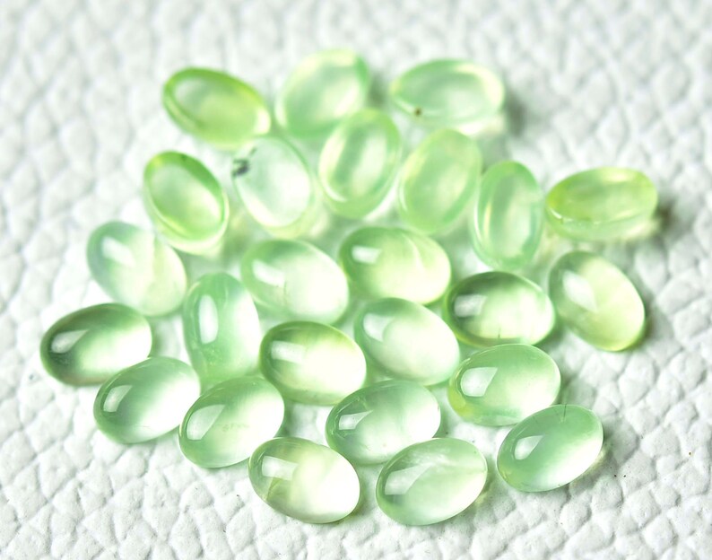 May include: A collection of oval-shaped, light green gemstones. The stones are smooth and polished, with a translucent appearance.
