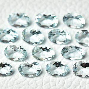 May include: A collection of 14 oval-shaped aquamarine gemstones, each with a light blue color and a faceted cut. The gemstones are arranged in a random pattern on a white surface.