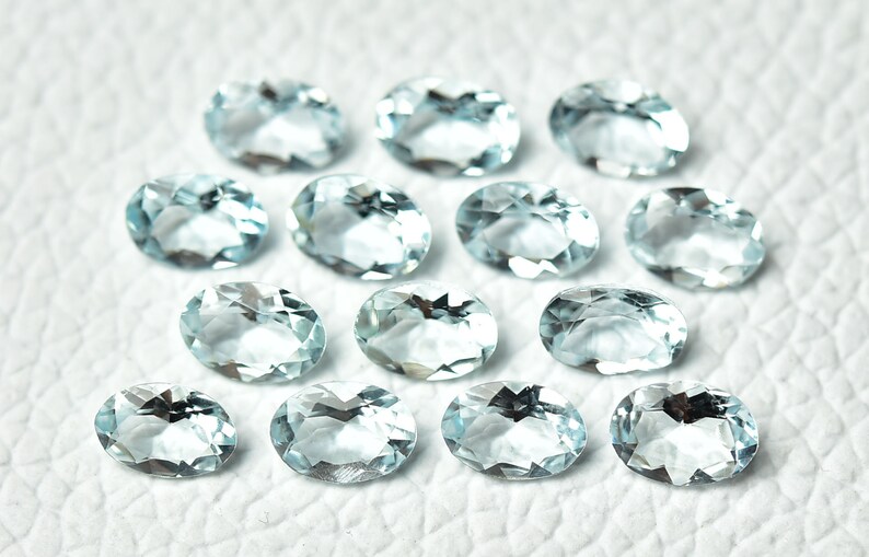 May include: A collection of 14 oval-shaped aquamarine gemstones, each with a light blue color and a faceted cut. The gemstones are arranged in a random pattern on a white surface.