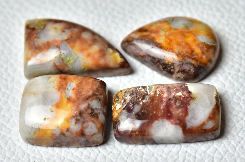 May include: Four polished, multi-colored agate cabochons with a white, brown, and orange color scheme. The cabochons are cut in different shapes, including rectangular and teardrop.