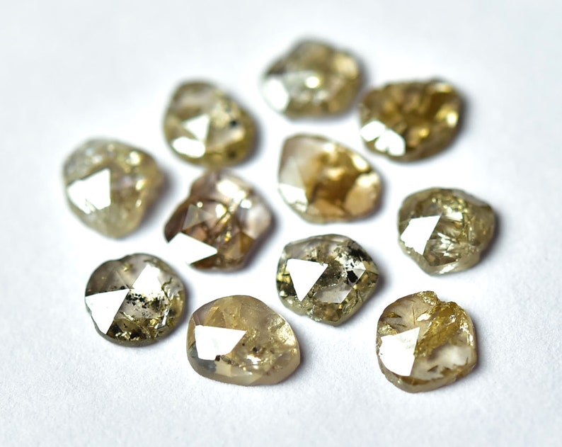 May include: A collection of ten faceted, rough-cut, light brown diamonds. The diamonds are arranged in a circle on a white surface.