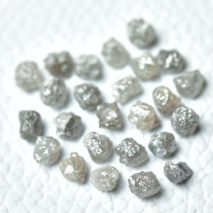 Puede incluir: Una colecci&oacute;n de diamantes grises en bruto, tambi&eacute;n conocidos como diamantes en bruto, esparcidos sobre una superficie blanca. Estos diamantes no est&aacute;n pulidos y tienen un aspecto natural y terroso.