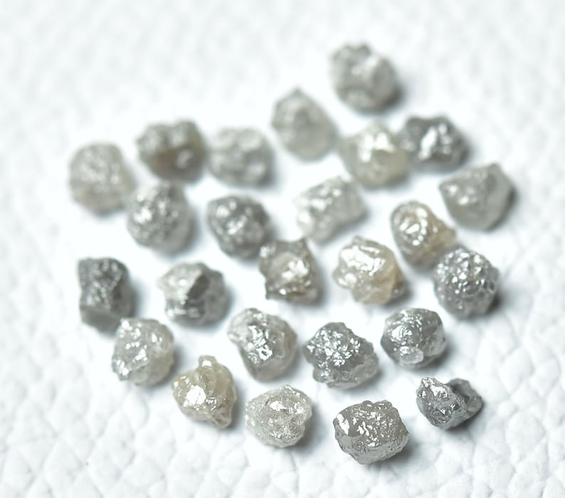 Puede incluir: Una colecci&oacute;n de diamantes grises en bruto, tambi&eacute;n conocidos como diamantes en bruto, esparcidos sobre una superficie blanca. Estos diamantes no est&aacute;n pulidos y tienen un aspecto natural y terroso.