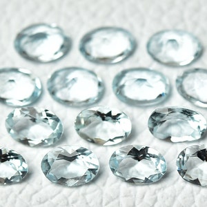 May include: A collection of oval-shaped aquamarine gemstones, each with a light blue hue and a smooth, polished finish. The stones are arranged on a white surface.