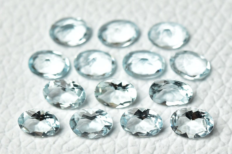 May include: A collection of oval-shaped aquamarine gemstones, each with a light blue hue and a smooth, polished finish. The stones are arranged on a white surface.