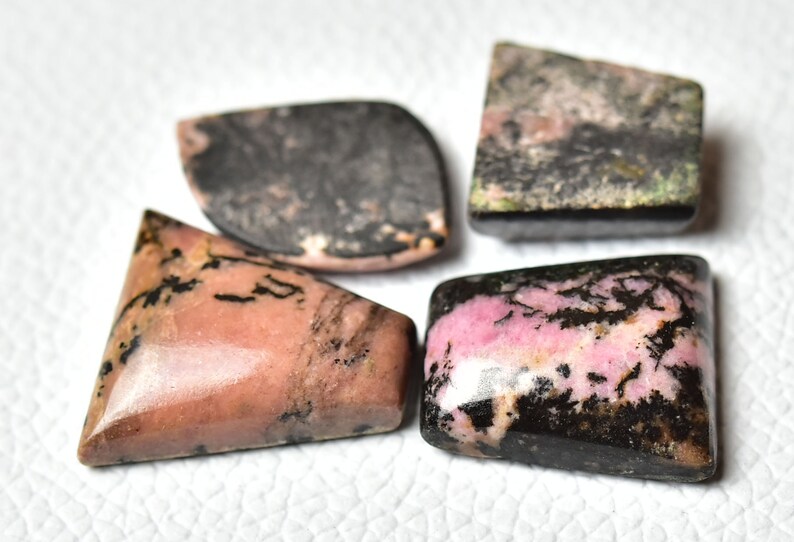 May include: Four cabochon cut gemstones with pink and black patterns. The stones are cut in different shapes, including a triangle, a rectangle, and two ovals.