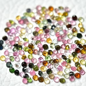 May include: A collection of round, faceted gemstones in various colours, including pink, green, blue, yellow, and brown. The gemstones are scattered on a white surface.