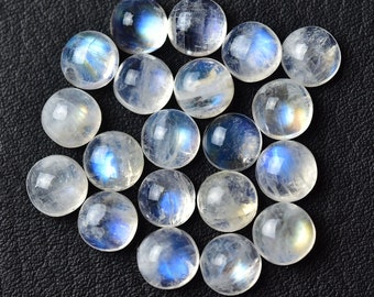 Puede incluir: Una colección de cabujones redondos de piedra lunar blanca con destellos iridiscentes azules y arcoíris. Las piedras son lisas y pulidas, y están dispuestas en un patrón aleatorio.