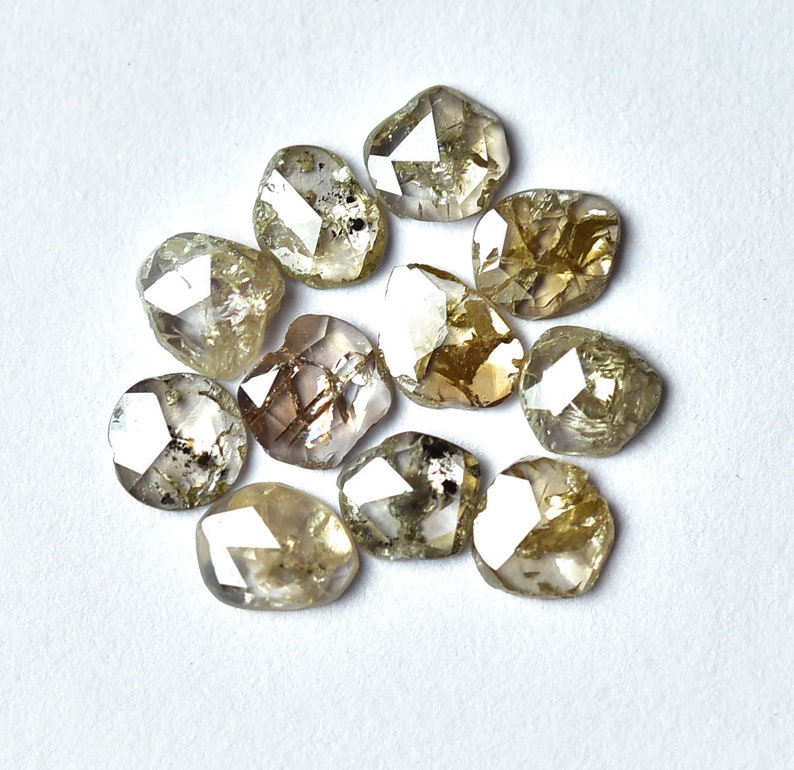 May include: A collection of twelve rough, faceted, natural diamonds. The diamonds are a light brown color with a translucent appearance. The diamonds are arranged in a circular pattern on a white surface.