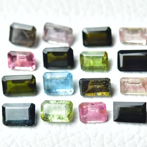 Op de afbeelding: Assortiment van 14 gefacetteerde edelstenen in verschillende kleuren, waaronder groen, roze, blauw, zwart en bruin. De edelstenen zijn in een rechthoekige vorm geslepen en zijn in rijen op een wit oppervlak gerangschikt.