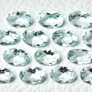 May include: A collection of 14 oval-shaped aquamarine gemstones, each with a light blue hue and a faceted cut. The stones are arranged in a random pattern on a white surface.