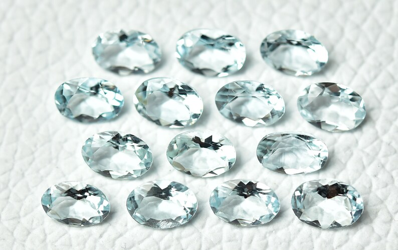 May include: A collection of 14 oval-shaped aquamarine gemstones, each with a light blue hue and a faceted cut. The stones are arranged in a random pattern on a white surface.