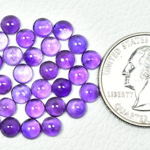 May include: A collection of 40 round, purple amethyst cabochons, each approximately 6mm in diameter. The cabochons are arranged in a loose pile and are shown next to a US quarter for size comparison.