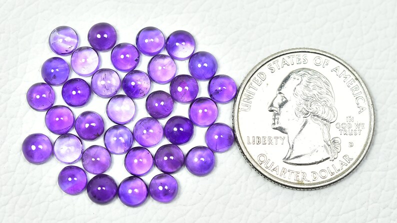 May include: A collection of 40 round, purple amethyst cabochons, each approximately 6mm in diameter. The cabochons are arranged in a loose pile and are shown next to a US quarter for size comparison.