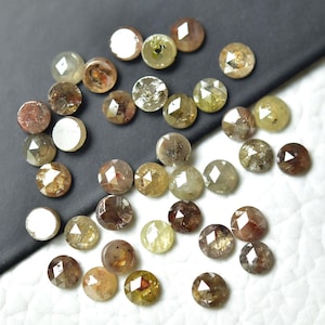 Op de afbeelding: Een verzameling ronde, gefacetteerde, ruwe diamanten in verschillende tinten bruin, grijs en groen. De diamanten zijn verspreid over een wit oppervlak.