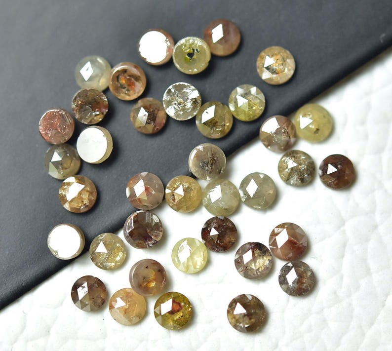 Op de afbeelding: Een verzameling ronde, gefacetteerde, ruwe diamanten in verschillende tinten bruin, grijs en groen. De diamanten zijn verspreid over een wit oppervlak.