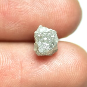 May include: A rough, grey diamond crystal with a faceted surface. The diamond is held in a hand.