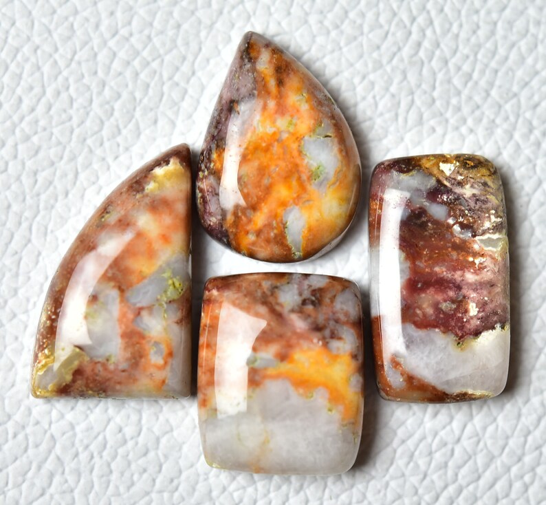 May include: Four cabochon cut gemstone cabochons in various shapes and sizes. The stones are a mix of white, brown, orange, and red with a marbled pattern.