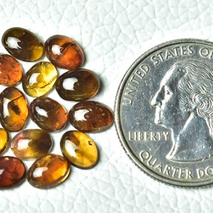 May include: A group of 15 oval-shaped, translucent, amber gemstones, each with a slightly different shade of brown and yellow. The gemstones are arranged in a loose pile and are about the size of a UK 20 pence coin.