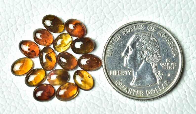May include: A group of 15 oval-shaped, translucent, amber gemstones, each with a slightly different shade of brown and yellow. The gemstones are arranged in a loose pile and are about the size of a UK 20 pence coin.