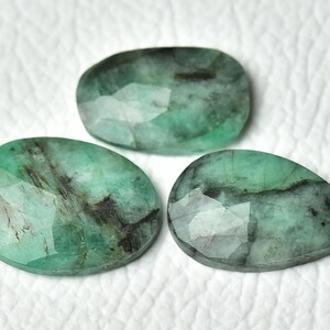 Könnte beinhalten: Drei facettierte Smaragd-Edelsteine in Grüntönen mit dunklen Einschlüssen. Die Edelsteine haben ovale, tropfenförmige und unregelmäßige Formen. Die Edelsteine werden auf einer weißen, strukturierten Oberfläche präsentiert.