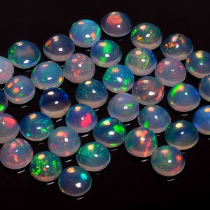 May include: A collection of round, white opal cabochons with iridescent rainbow flashes. The opals are arranged in a loose pile on a black background.