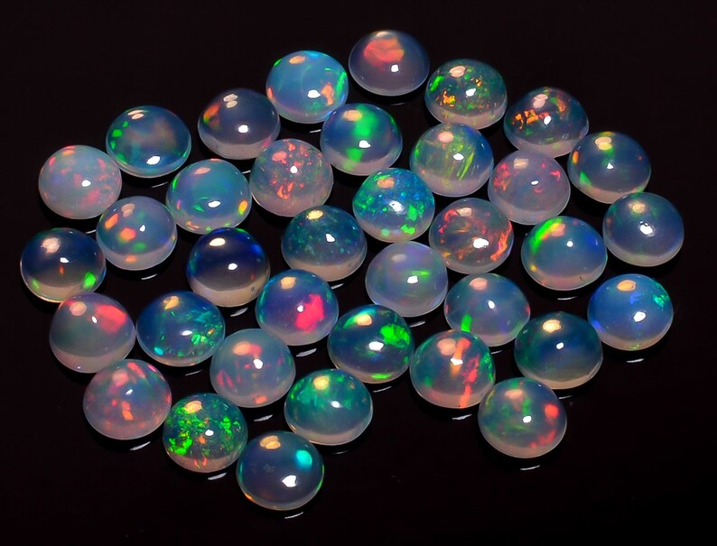 May include: A collection of round, white opal cabochons with iridescent rainbow flashes. The opals are arranged in a loose pile on a black background.