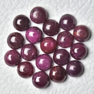 May include: A collection of polished, round gemstones in varying shades of deep red and purple. The gems have a smooth, reflective surface, with natural inclusions visible. The arrangement is on a white, textured surface, showcasing the gems' rich colors and luster.