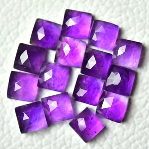 May include: A collection of 14 faceted amethyst gemstones, each cut into a square shape with rounded corners. The stones are a deep purple colour and have a glossy finish.