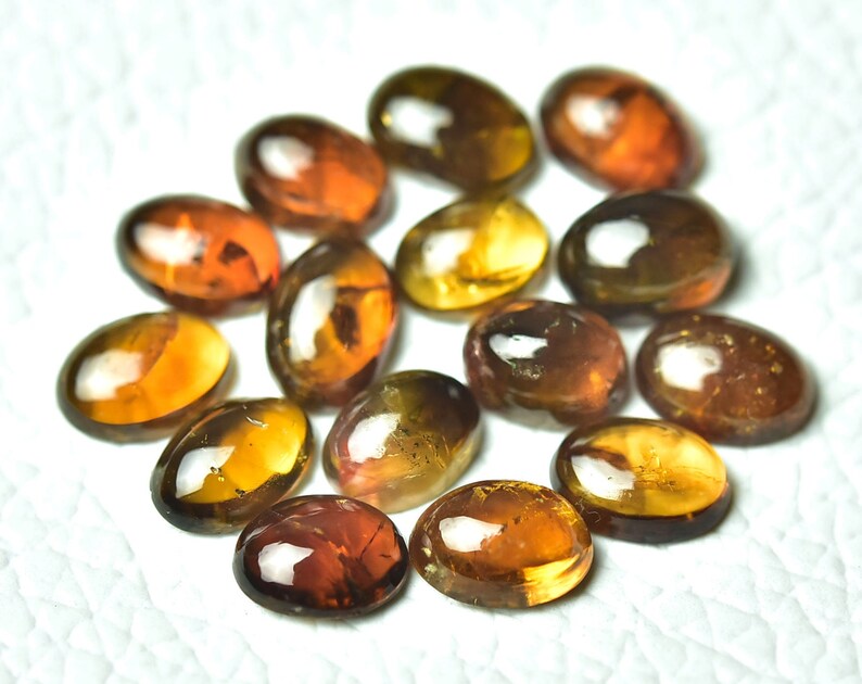 May include: A collection of 14 oval-shaped gemstones in shades of brown, orange, and yellow. The gemstones are smooth and polished, with a glossy finish.