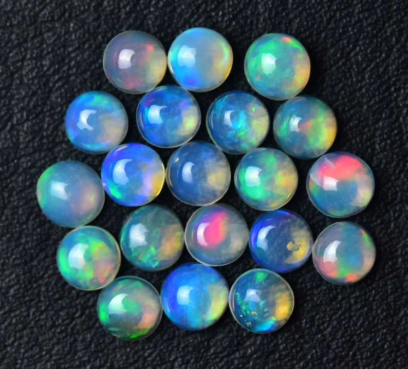May include: A collection of 20 round opal cabochons in various shades of blue, green, and white. The cabochons are arranged in a circular pattern on a black surface.