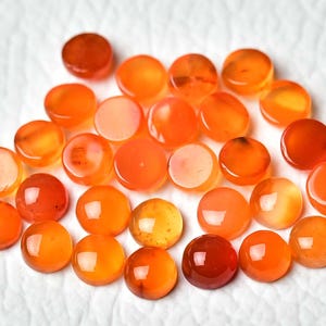 Pu&ograve; includere: Una collezione di pietre preziose rotonde e lucide in tonalit&agrave; arancioni e rosse. Le pietre variano in intensit&agrave; di colore, dall'arancione chiaro al marrone rossastro scuro, e sono disposte su una superficie bianca e strutturata. Queste pietre preziose sono probabilmente utilizzate per la creazione di gioielli.