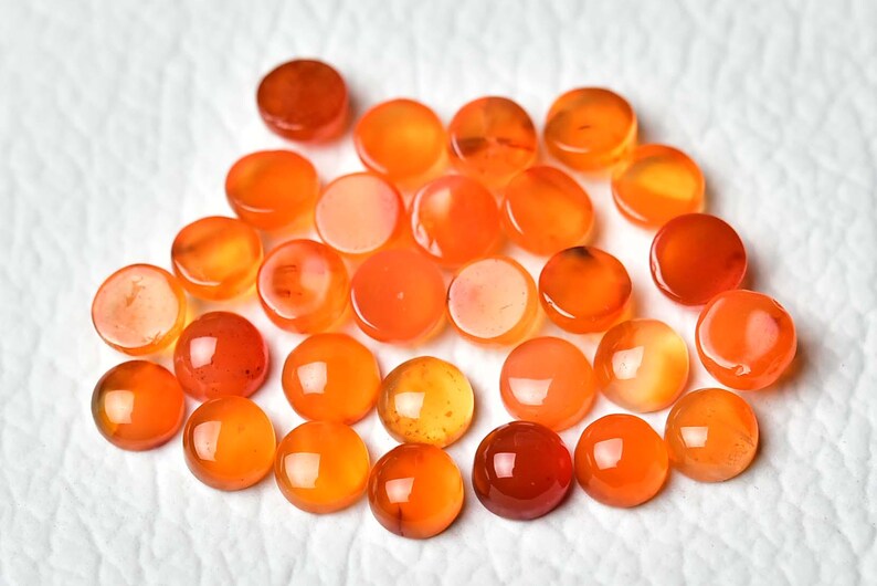 Pu&ograve; includere: Una collezione di pietre preziose rotonde e lucide in tonalit&agrave; arancioni e rosse. Le pietre variano in intensit&agrave; di colore, dall'arancione chiaro al marrone rossastro scuro, e sono disposte su una superficie bianca e strutturata. Queste pietre preziose sono probabilmente utilizzate per la creazione di gioielli.