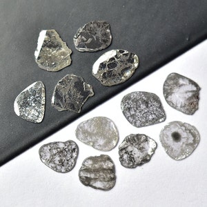 Op de afbeelding: Een verzameling van 11 ruwe, ongeslepen, grijze diamanten. De diamanten zijn van verschillende vormen en maten. Sommige diamanten hebben een zilveren glans.