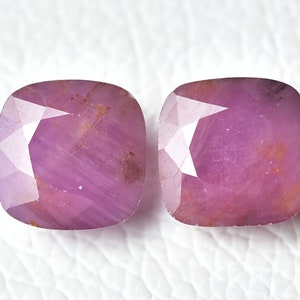 May include: Two pink cushion-cut gemstones with a faceted finish. The gemstones have a slightly reddish hue and are set against a white background.