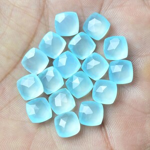 K&ouml;nnte beinhalten: Eine Sammlung von 16 hellblauen Edelstein-Cabochons, jeder mit einer facettierten quadratischen Form. Die Steine sind in einem lockeren Haufen auf einer Hand angeordnet.