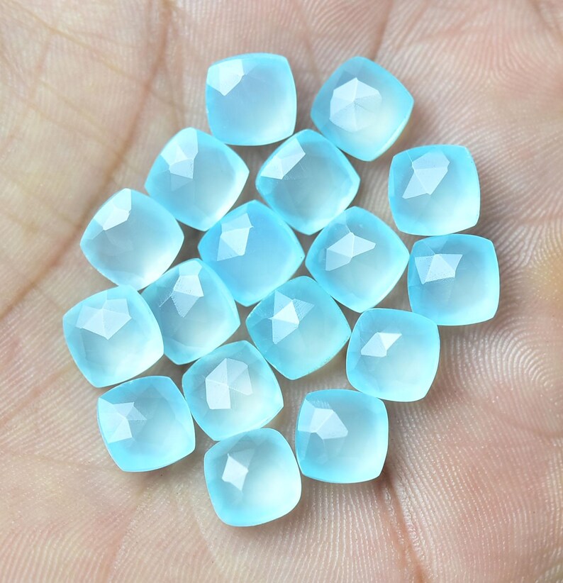 K&ouml;nnte beinhalten: Eine Sammlung von 16 hellblauen Edelstein-Cabochons, jeder mit einer facettierten quadratischen Form. Die Steine sind in einem lockeren Haufen auf einer Hand angeordnet.