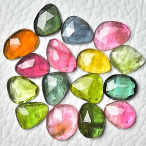 Op de afbeelding: Een verzameling gefacetteerde edelstenen in verschillende kleuren, waaronder roze, groen, geel, oranje en blauw. De edelstenen zijn onregelmatig van vorm en lijken gepolijst. De achtergrond is een getextureerd wit oppervlak.