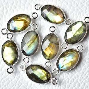 Op de afbeelding: Acht ovale labradorit edelsteen kralen met zilverkleurige springringen. De kralen zijn een mix van groene, gouden en blauwe iriserende kleuren.