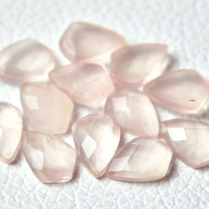 May include: A collection of pale pink rose quartz gemstones, each cut into a faceted, teardrop shape. The stones are arranged on a white surface.