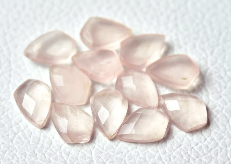 May include: A collection of pale pink rose quartz gemstones, each cut into a faceted, teardrop shape. The stones are arranged on a white surface.