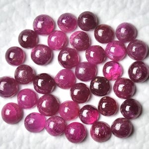 以下が含まれることがあります： 赤とピンクの色合いが異なる、丸く磨かれたルビーの宝石のコレクション。宝石は白い表面に散らばっており、自然な色と光沢を示しています。宝石はジュエリー作りに適しています。