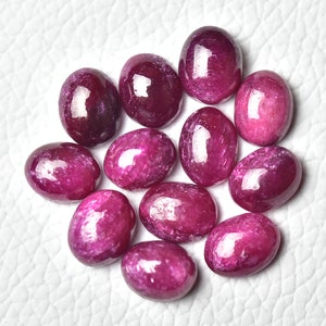 May include: A collection of 12 oval-shaped, pink ruby gemstones. The gemstones are smooth and polished, with a slight shimmer.