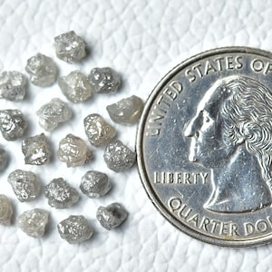Puede incluir: Una colecci&oacute;n de peque&ntilde;os diamantes grises rugosos, algunos con un brillo plateado, est&aacute;n dispersos sobre una superficie blanca. Se incluye una moneda de un cuarto de d&oacute;lar estadounidense para la escala.