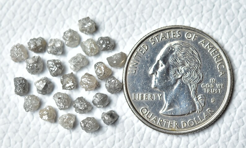 Puede incluir: Una colecci&oacute;n de peque&ntilde;os diamantes grises rugosos, algunos con un brillo plateado, est&aacute;n dispersos sobre una superficie blanca. Se incluye una moneda de un cuarto de d&oacute;lar estadounidense para la escala.