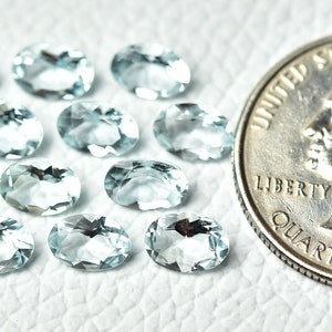 May include: A collection of 14 oval-shaped aquamarine gemstones, ranging in size from small to medium. The stones are a light blue color and have a smooth, polished finish. A US quarter is included for size reference.