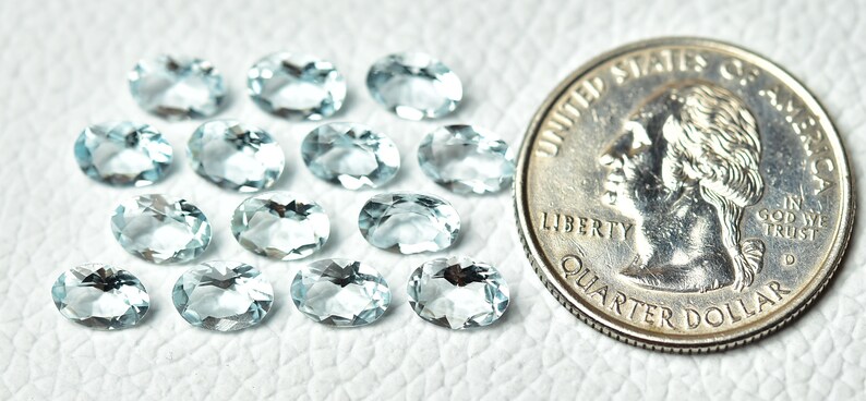 May include: A collection of 14 oval-shaped aquamarine gemstones, ranging in size from small to medium. The stones are a light blue color and have a smooth, polished finish. A US quarter is included for size reference.